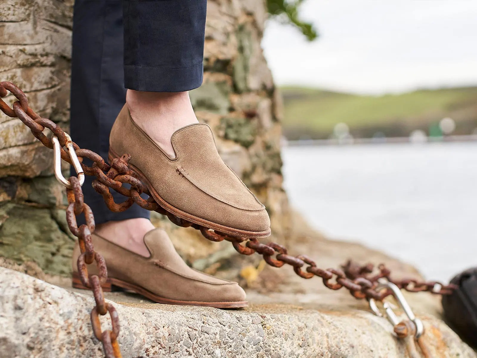 A model wearing a pair of Crockett & Jones loafers. One foot is resting on a rusty chain.