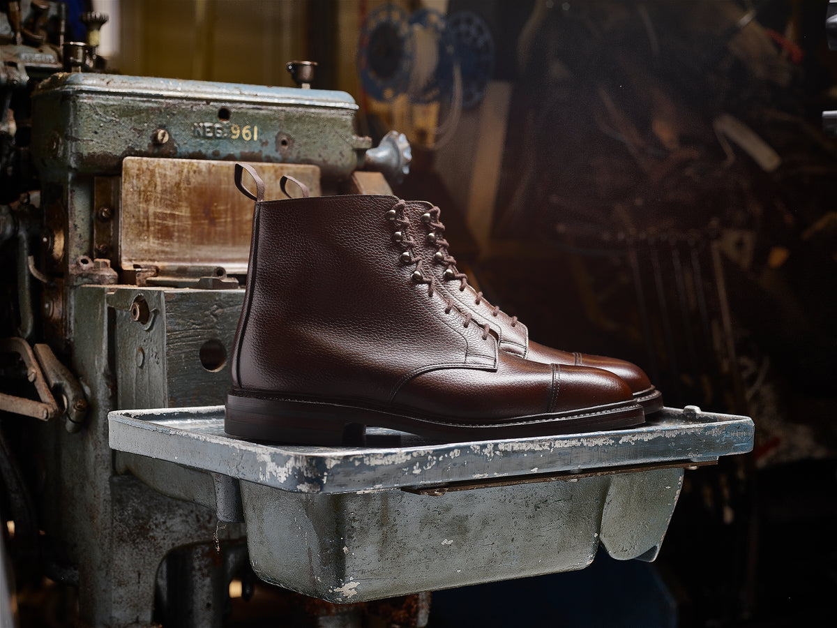 A dark brown leather lace-up ankle boot with a cap toe, displayed on a weathered metal workbench in a workshop setting.