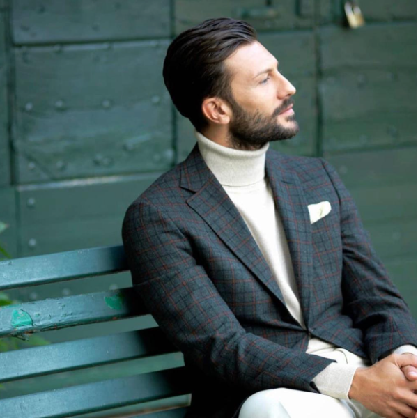 A man in a checked blazer and rollneck sweater is sitting on a bench.